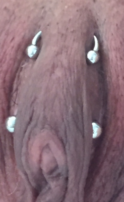 An ornamental Horizontal Clitoral Hood (HCH) piercing above (and a triangle piercing below) with open circular barbells "U-bars"